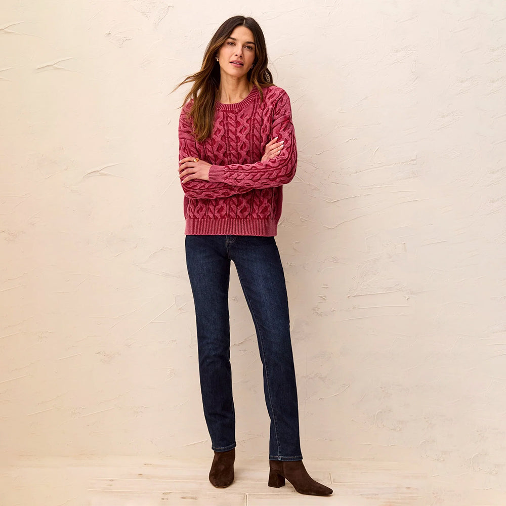 A woman modeling a dusty rose cable-knit crewneck sweater, paired with dark blue jeans and brown suede ankle boots, standing against a textured off-white wall.