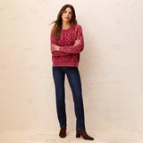 A woman modeling a dusty rose cable-knit crewneck sweater, paired with dark blue jeans and brown suede ankle boots, standing against a textured off-white wall.
