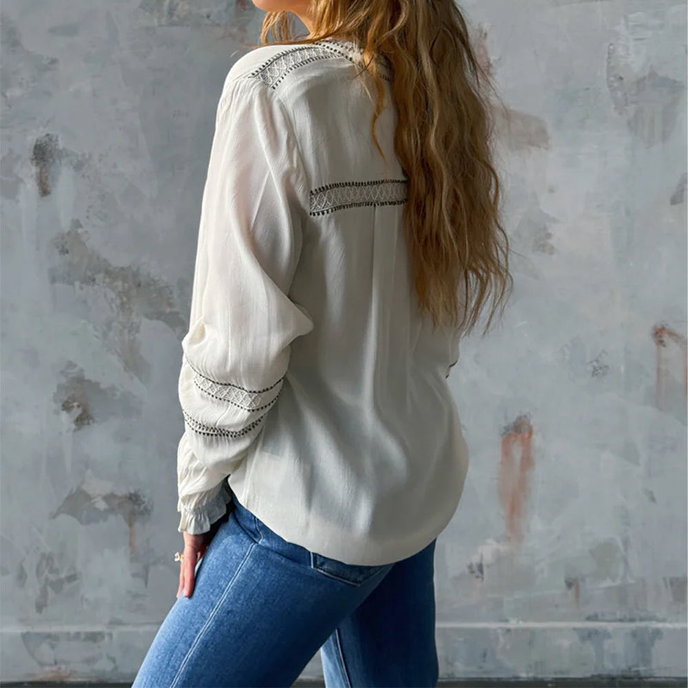 Person wearing a white blouse with lace details and blue jeans against a textured wall.
