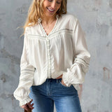 Woman wearing a white blouse with lace details and blue jeans against a gray wall.