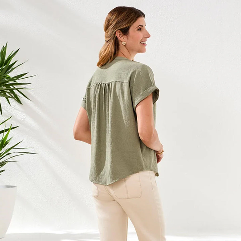 Woman wearing a green blouse and white pants standing against a white background with plants.