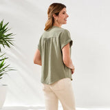 Woman wearing a green blouse and white pants standing against a white background with plants.