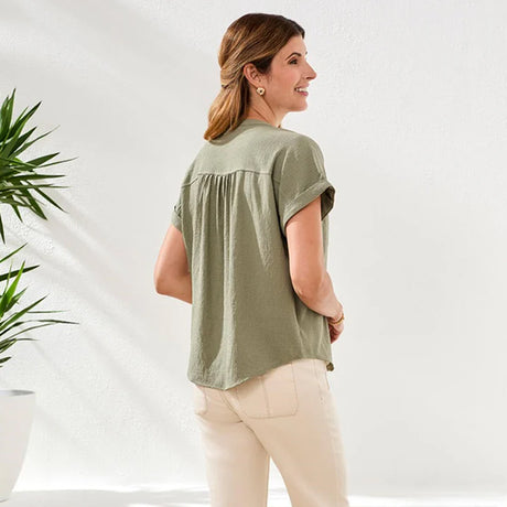 Woman wearing a green blouse and white pants standing against a white background with plants.