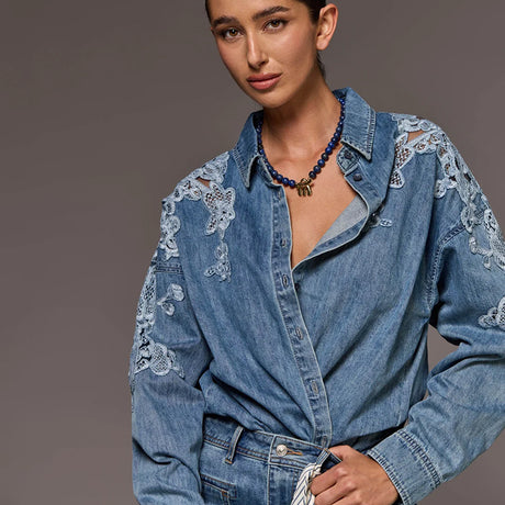 A woman wearing a light blue denim shirt with intricate white floral lace embroidery on the sleeves, styled with a blue beaded necklace against a solid gray studio background.