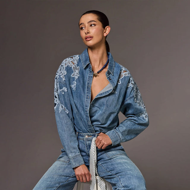 A model wearing a blue denim jumpsuit with white floral embroidery on the sleeves, paired with a matching blue denim belt and a striped tie, posed against a solid dark gray studio background.