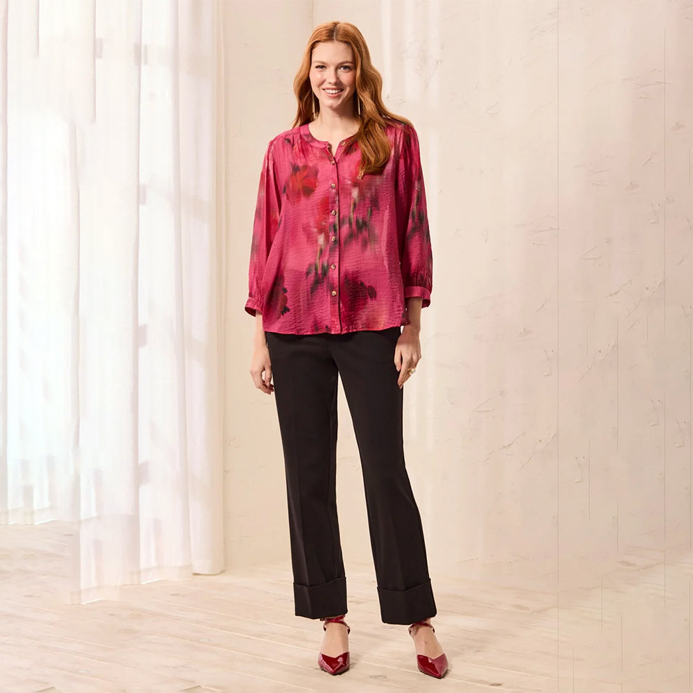 A woman stands in a bright indoor setting wearing a pink floral long-sleeve blouse, black pants, and red pointed heels.