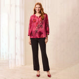 A woman stands in a bright indoor setting wearing a pink floral long-sleeve blouse, black pants, and red pointed heels.
