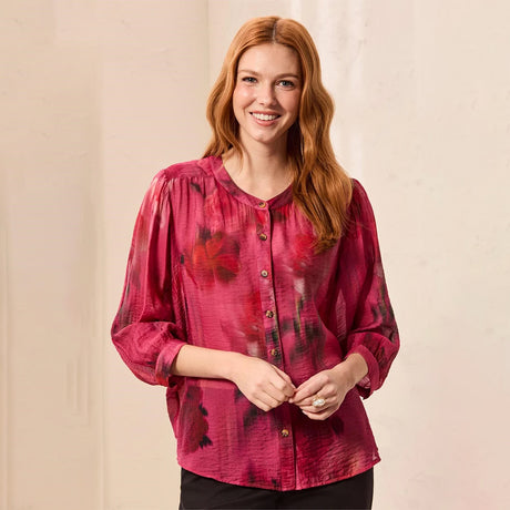 A smiling woman with long wavy red hair wears a long-sleeve button-front blouse in a deep magenta pink with dark red and black floral patterns. She is standing against a neutral, light beige textured wall.