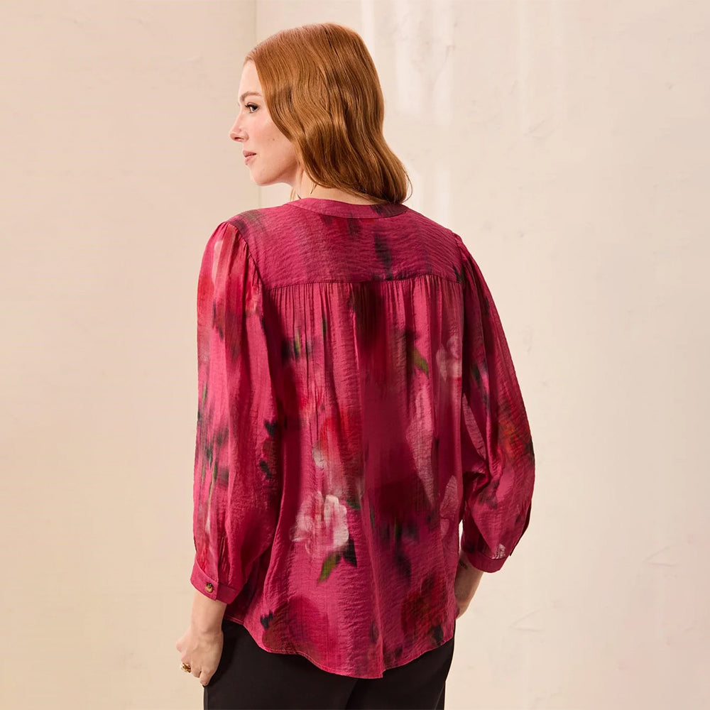 A woman with red hair is shown from the back wearing a sheer, textured magenta pink blouse with a dark floral pattern and black pants, standing against a light beige wall.