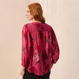 A woman with red hair is shown from the back wearing a sheer, textured magenta pink blouse with a dark floral pattern and black pants, standing against a light beige wall.