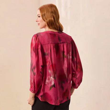 A woman with red hair is shown from the back wearing a sheer, textured magenta pink blouse with a dark floral pattern and black pants, standing against a light beige wall.