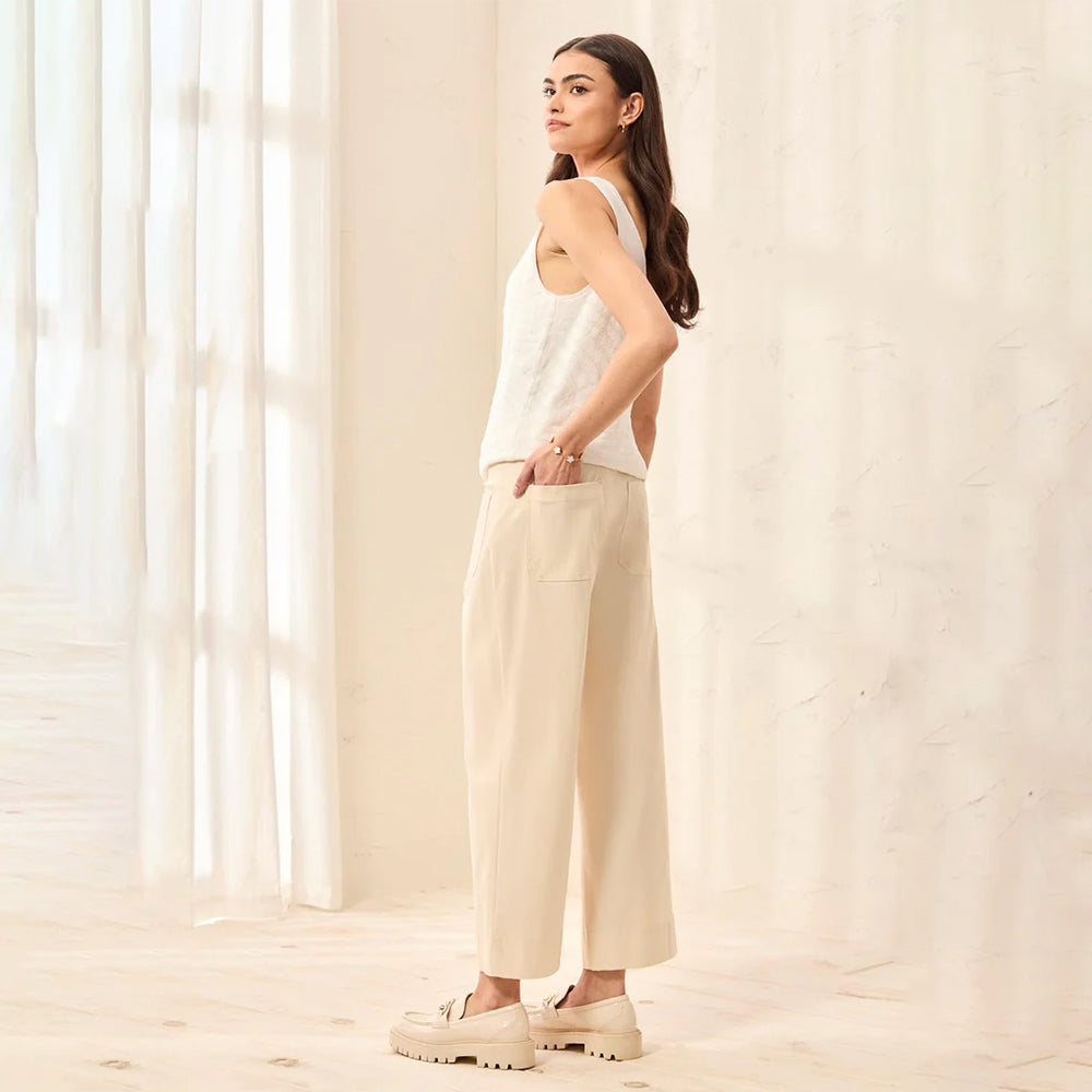Woman wearing a white sleeveless top and beige pants standing in a softly lit room.