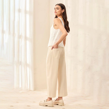 Woman wearing a white sleeveless top and beige pants standing in a softly lit room.