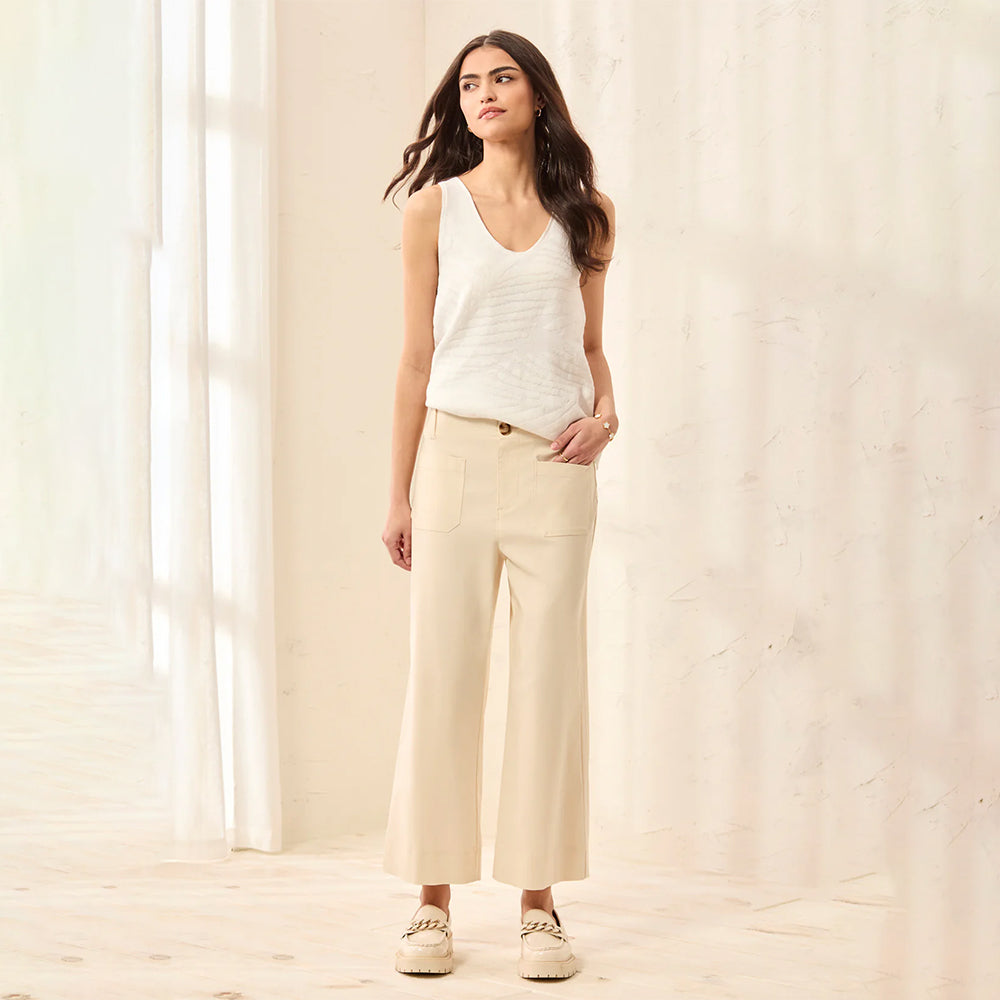 Woman wearing a white sleeveless top and beige pants standing in a softly lit room.