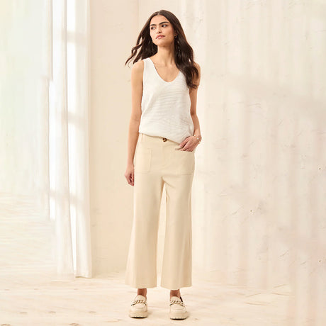 Woman wearing a white sleeveless top and beige pants standing in a softly lit room.