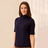 A woman wearing a short-sleeve mock neck top in dark navy blue, standing against a light beige textured wall.