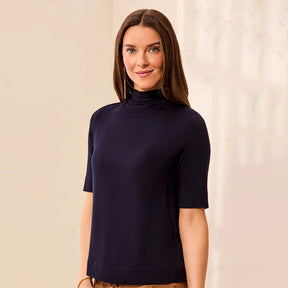 A woman wearing a short-sleeve mock neck top in dark navy blue, standing against a light beige textured wall.