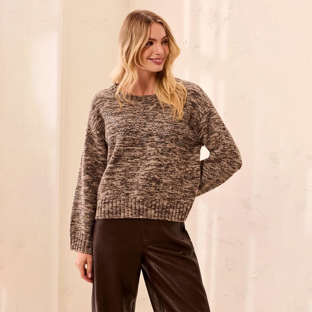 A woman modeling a long-sleeve crewneck knit sweater with a brown and beige marled pattern, paired with dark brown pants, set against a neutral cream-colored wall.