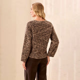 A woman shown from the back wearing a marled brown and beige knit sweater with long sleeves, paired with dark brown pants, standing against a textured off-white wall.