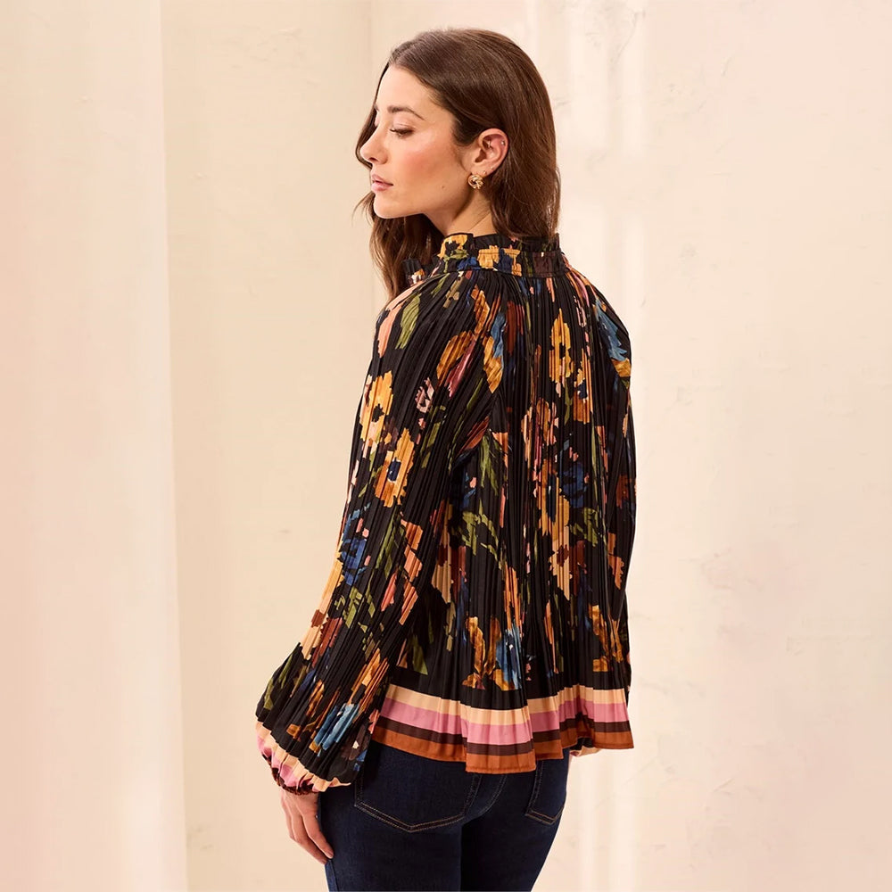 A woman wearing a black pleated long-sleeve blouse with a colorful floral and abstract print, paired with dark blue jeans, standing against a light beige wall.