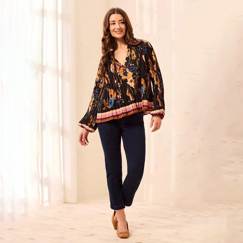 A woman modeling a black long-sleeve blouse with a vibrant orange, yellow, and blue floral pattern, paired with dark blue jeans and tan heels, standing in a brightly lit room with white curtains.