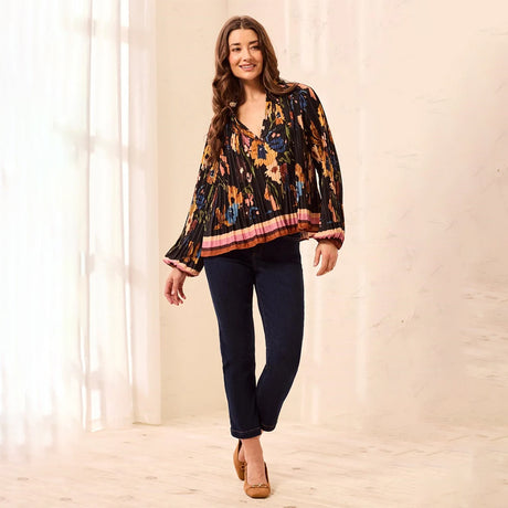 A woman modeling a black long-sleeve blouse with a vibrant orange, yellow, and blue floral pattern, paired with dark blue jeans and tan heels, standing in a brightly lit room with white curtains.