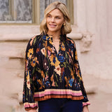 A woman wearing a black pleated long-sleeve blouse with a colorful floral print and a striped hem, paired with dark blue jeans, standing outdoors against a light beige stone wall.