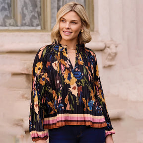 A woman wearing a black pleated long-sleeve blouse with a colorful floral print and a striped hem, paired with dark blue jeans, standing outdoors against a light beige stone wall.