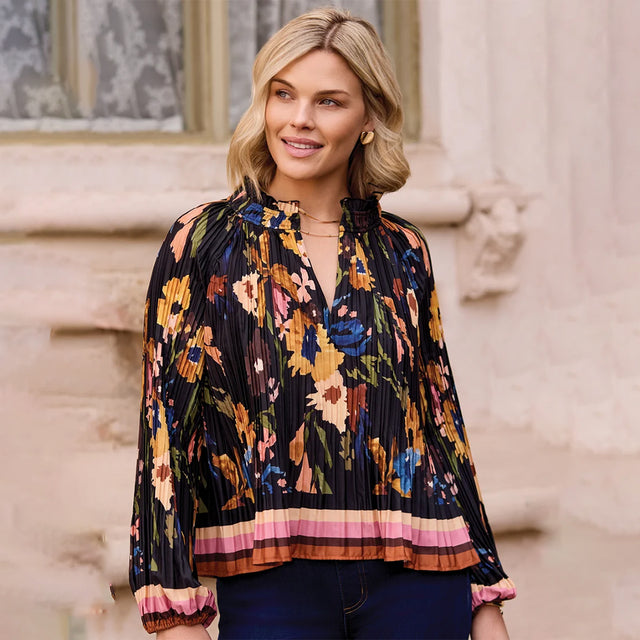 A woman wearing a black pleated long-sleeve blouse with a colorful floral print and a striped hem, paired with dark blue jeans, standing outdoors against a light beige stone wall.