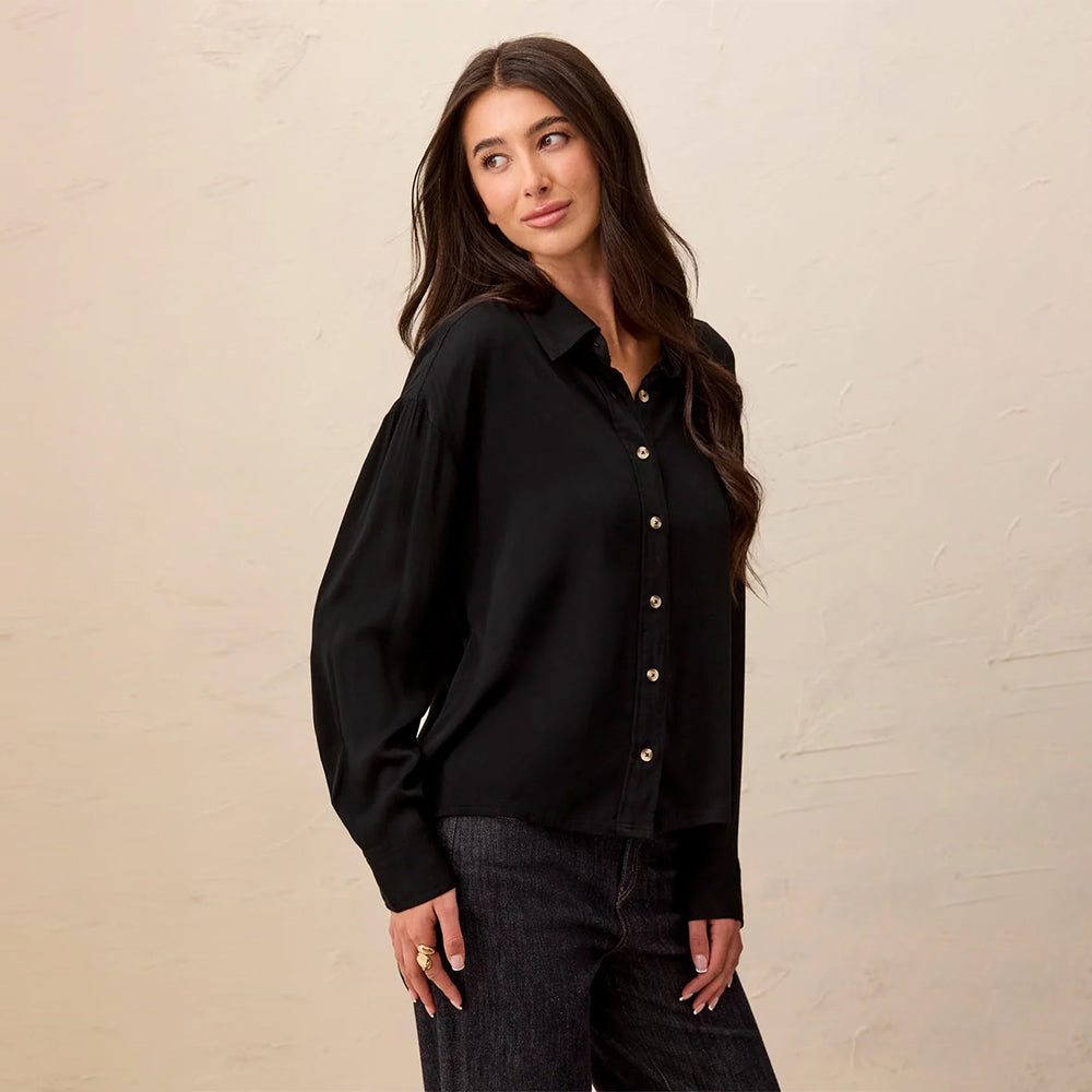 A woman wearing a black long-sleeve button-up blouse with gold-tone buttons, paired with dark jeans, standing against a textured beige wall.