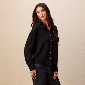 A woman wearing a black long-sleeve button-up blouse with gold-tone buttons, paired with dark jeans, standing against a textured beige wall.