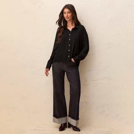 A woman stands against a textured off-white wall wearing a black long-sleeve button-front blouse with dark wide-leg jeans featuring rolled gray cuffs and black shoes.