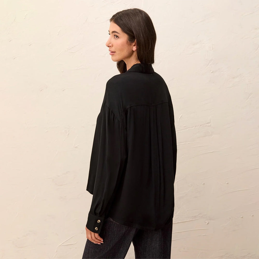 A woman wearing a black long-sleeve blouse with pleated detailing on the back, shown from a side-back angle against a textured off-white wall.