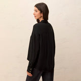 A woman wearing a black long-sleeve blouse with pleated detailing on the back, shown from a side-back angle against a textured off-white wall.