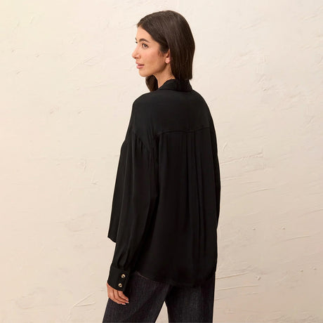 A woman wearing a black long-sleeve blouse with pleated detailing on the back, shown from a side-back angle against a textured off-white wall.