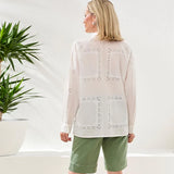 Woman wearing a light-colored blouse with intricate patterns and green shorts in a minimalistic setting.