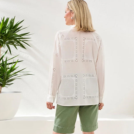 Woman wearing a light-colored blouse with intricate patterns and green shorts in a minimalistic setting.