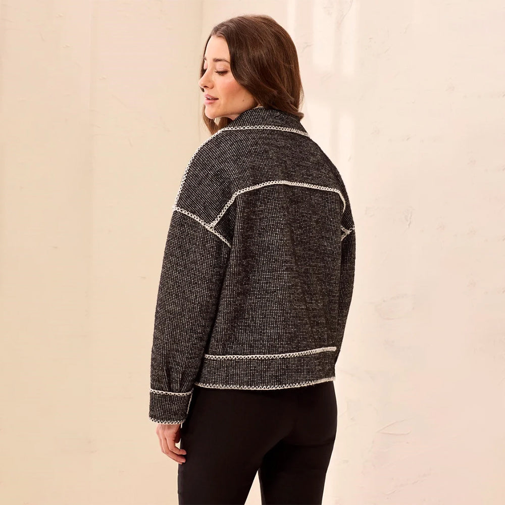 A woman wearing a dark gray textured knit cardigan with white contrast stitching, shown from a side-back angle against a warm beige wall.