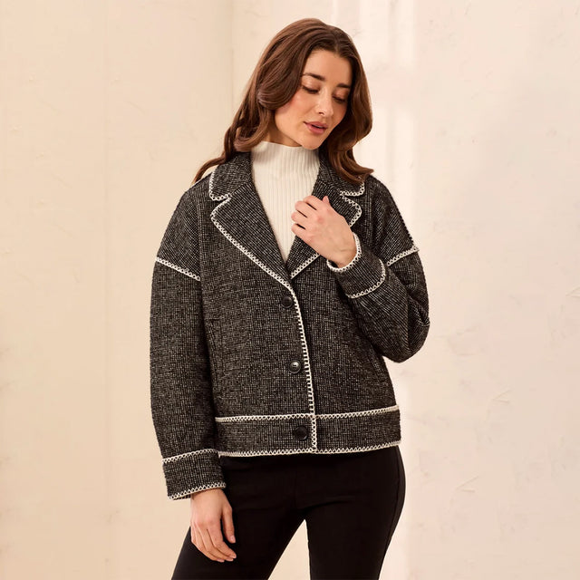 A woman wearing a dark gray and black tweed blazer with white contrast stitching, styled over a white ribbed turtleneck and black pants, standing against a light beige textured wall.