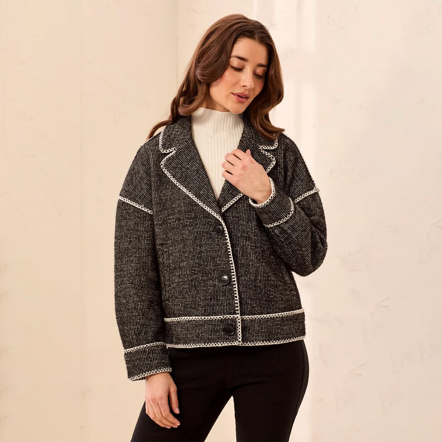 A woman wearing a dark gray and black tweed blazer with white contrast stitching, styled over a white ribbed turtleneck and black pants, standing against a light beige textured wall.