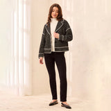 A woman modeling a dark gray and black tweed jacket with white piping, worn over a white turtleneck and black pants, standing in a brightly lit room with white curtains.