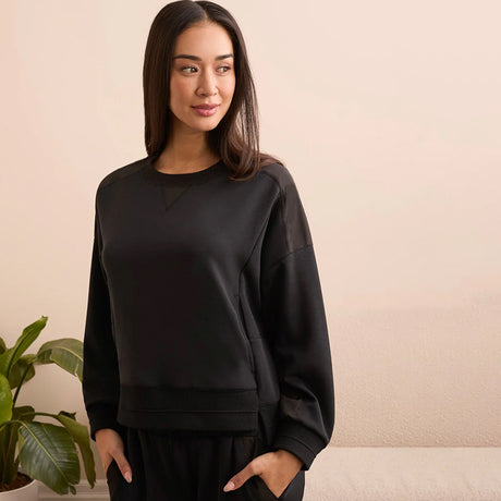 Woman wearing a black sweatshirt standing against a beige wall with a plant to the left.
