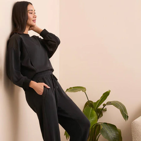 Woman in black outfit leaning against a wall with a plant nearby