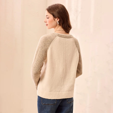 A woman shown from the back wearing a cream and taupe knit raglan sweater with blue jeans, standing against a soft beige backdrop.