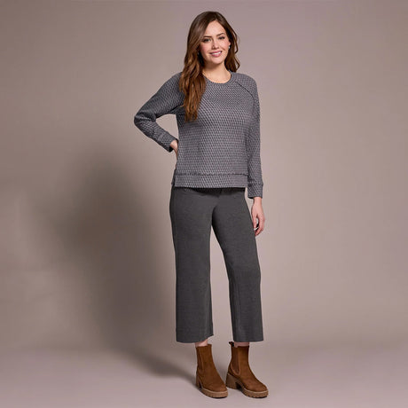 Woman wearing a gray patterned top and pants with brown boots against a beige background