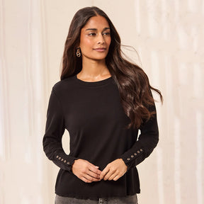 A woman modeling a black long-sleeve top with decorative gold-tone buttons along the cuffs, set against a soft beige curtain background.