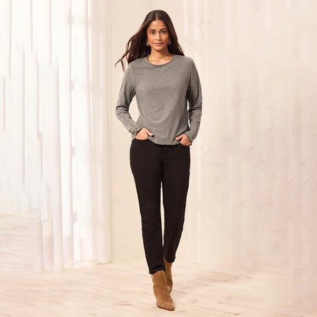 A woman modeling a gray long-sleeve crewneck sweater, black pants, and tan ankle boots in a bright, modern indoor setting with white curtains.