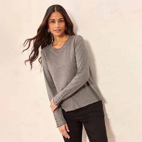 A woman modeling a heather gray long-sleeve knit top with button cuffs, paired with black pants, standing against a textured off-white wall.