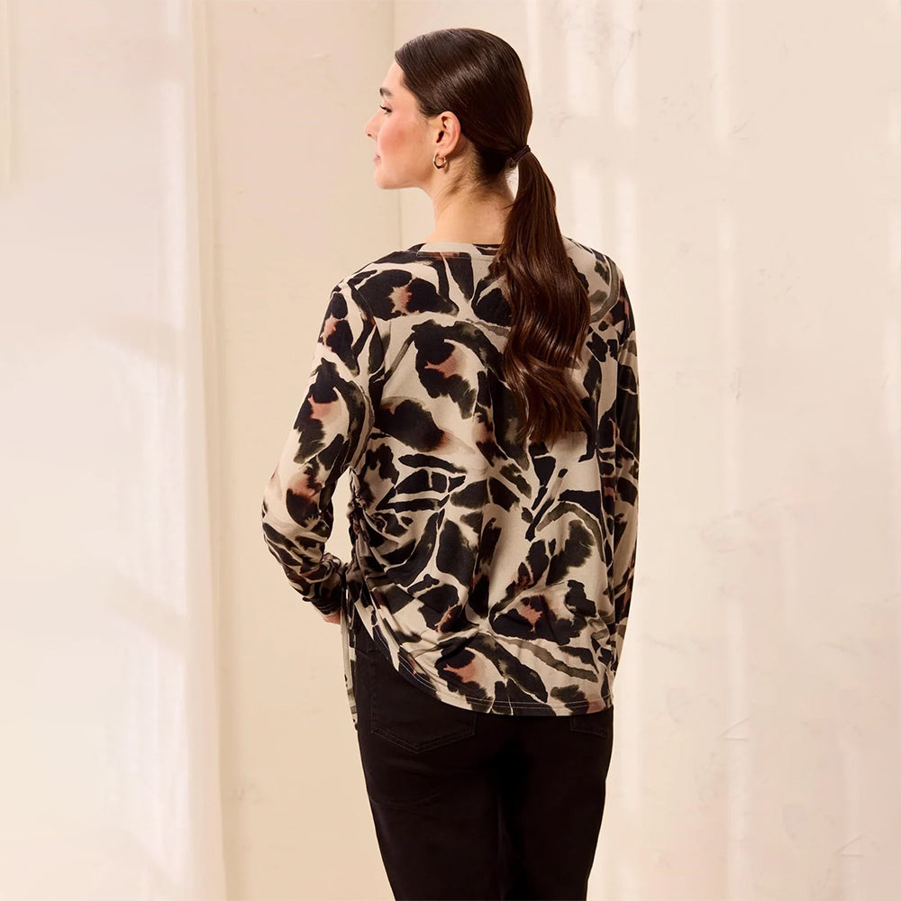 A woman shown from the back wearing a long-sleeve blouse with a black, beige, and brown abstract floral pattern, paired with black pants, set against a warm off-white background.