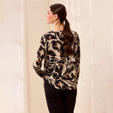 A woman shown from the back wearing a long-sleeve blouse with a black, beige, and brown abstract floral pattern, paired with black pants, set against a warm off-white background.